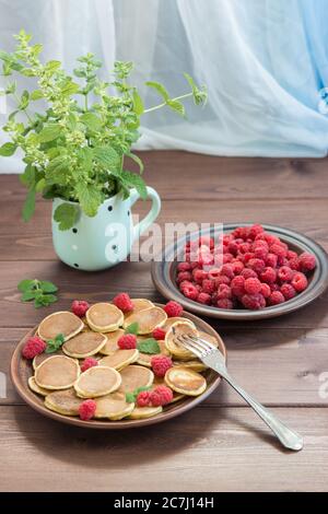 Crêpe de céréales sur la fourchette. Framboises fraîches mûres rouges dans une assiette brune et un bouquet de Melissa dans une tasse sur la table en bois sombre près de la fenêtre. Suppr Banque D'Images