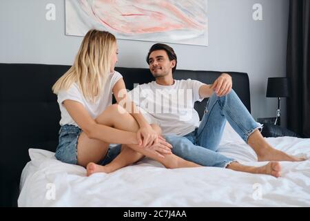 Jeune homme de brunette beau regardant de façon rêveuse une jolie femme blonde assise sur le lit ensemble dans un hôtel moderne et confortable Banque D'Images