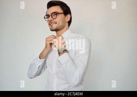 Jeune homme souriant brunes en chemise blanche et lunettes avec montre à main regardant de côté sur fond blanc seul Banque D'Images