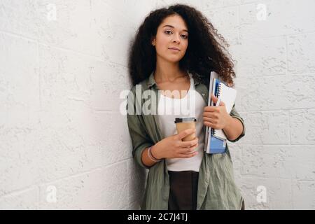 Jeune fille étudiante attrayante avec cheveux en forme de boucles sombres dans une chemise kaki tenant un ordinateur portable avec bloc-notes et une tasse de café pour aller dans les mains regardant dans Banque D'Images