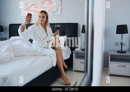 Jeune femme blonde souriante, peignoir blanc tenant un verre de vin rouge à la main, prenant volontiers des photos sur un téléphone portable assis sur le lit dans un hôtel moderne Banque D'Images