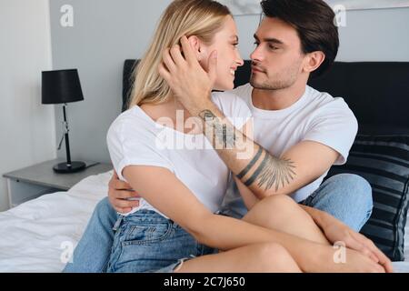 Beau brunette homme rêveur lissant les cheveux et embrassant jolie femme blonde souriante. Jeune beau couple en T-shirts blancs assis sur le lit Banque D'Images