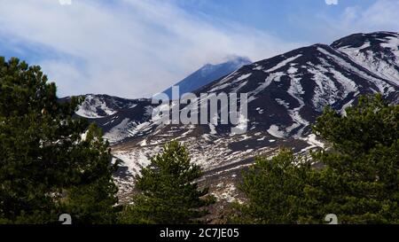 Etna, Parc National de l'Etna, Parco dell – Etna, sommet de l'Etna derrière les nuages en arrière-plan, sommets enneigés au milieu, arbres verts en premier plan Banque D'Images