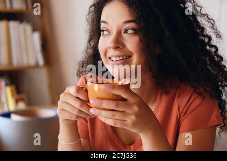 Portrait d'une jeune femme souriante et attrayante avec des cheveux en courbure foncé tenant une tasse de café orange dans les mains, regardant avec joie la maison moderne et confortable seule Banque D'Images