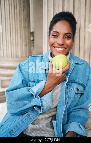 Belle fille afro-américaine joyeuse en veste denim tenant la pomme joyeusement regarder dans l'appareil photo extérieur Banque D'Images