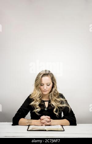 Femme blonde lisant une bible sur la table contre un arrière-plan blanc Banque D'Images