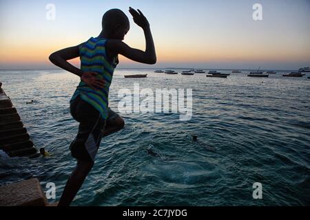 Un garçon non identifié saute dans la mer comme le soleil descend dans la vieille ville en pierre de Zanzibar, Zanzibar, Tanzanie, l'île a une longue histoire avec le commerce des esclaves et a été dirigé par les Arabes omanais au début du XIXe siècle, L'île est majoritairement musulmane et est devenue une destination touristique populaire ces dernières années, l'île est une promenade en bateau de deux heures de Dar es Salaam, Tanzanie sur la côte est africaine, l'île fait partie de la Tanzanie, Banque D'Images