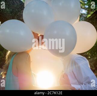 Couple embrassant couvert de ballons blancs dans un jardin sous la lumière du soleil Banque D'Images