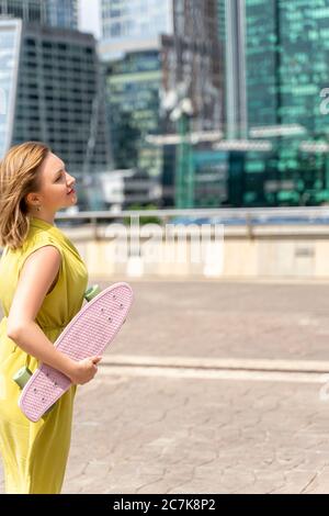 la 30ème belle femme aux cheveux rouges avec un skateboard regarde de rêve les gratte-ciels d'une grande ville Banque D'Images