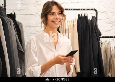 Jeune jolie femme joyeuse utilisant le téléphone portable travaillant dans la salle d'exposition de mode Banque D'Images