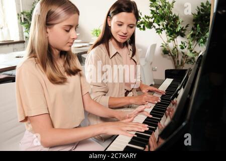 Une jeune fille adolescente aux cheveux longs et blonds, assise au piano près de sa mère, tout en jouant du matériel musical dans le salon Banque D'Images