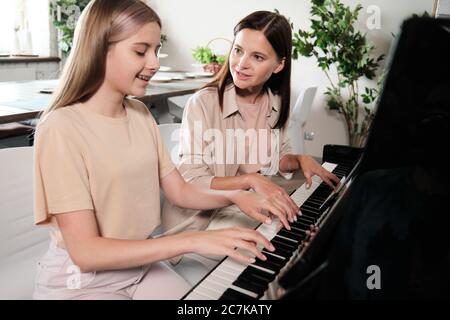 Jeune femme brune consultant sa fille adolescente intelligente, assise au piano et jouant ensemble dans un environnement familial Banque D'Images