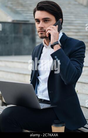 Jeune homme de brunette à barbe en chemise blanche et costume classique assis sur le banc avec ordinateur portable sur les genoux, regardant de côté parler sur le téléphone portable Banque D'Images