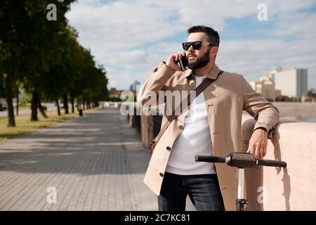Jeune agent élégant avec scooter électrique parlant sur téléphone mobile en milieu urbain tout en se tenant entre le bord de la rivière et le parc Banque D'Images