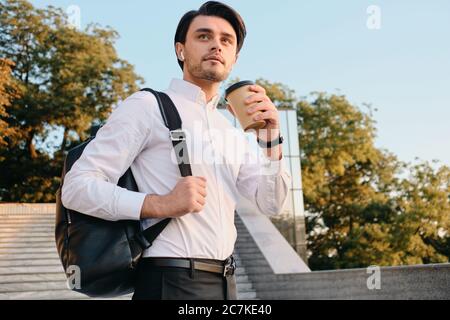 Jeune homme de brunette à barbe en chemise blanche avec écouteurs sans fil debout avec une tasse de café à la main et sac à dos sur l'épaule Banque D'Images