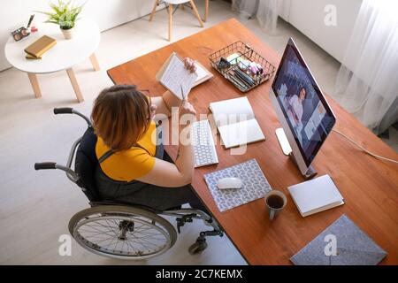Jeune désactiver enseignante de consultation étudiante en ligne et expliquant des notes dans le copybook tout en étant assise en fauteuil roulant par ordinateur Banque D'Images