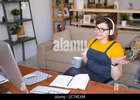 Jeune femme moderne désactiver dans le casque d'écoute prendre un verre et parler avec le professeur pendant la leçon en ligne devant l'ordinateur Banque D'Images