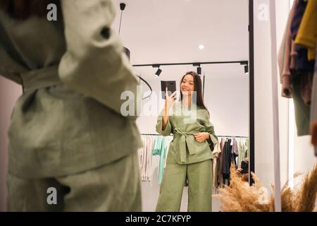 Belle fille asiatique essayant de costume élégant prenant heureusement selfie dans le miroir dans le dressing de magasin de vêtements Banque D'Images