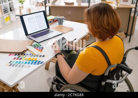 Jeune designer contemporain en fauteuil roulant utilisant une tablette avec stylet tout en travaillant sur les nouvelles technologies de communication devant un ordinateur portable Banque D'Images