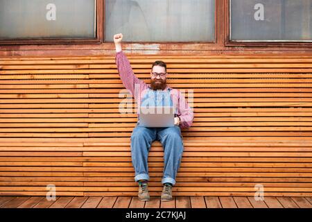 Gagnant. Portrait d'un boxeur hippie gai et brutal dans une combinaison bleue, chemise à carreaux travaillant sur un ordinateur portable assis sur un banc levant la main en fête Banque D'Images