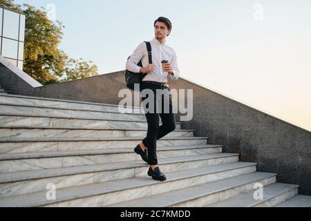 Jeune homme de brunette à barbe en chemise blanche avec écouteurs sans fil tenant une tasse de café à la main et sac à dos sur l'épaule judicieusement Banque D'Images