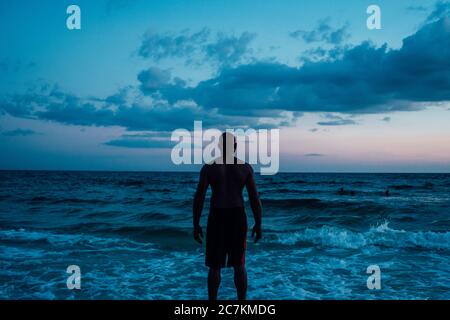 Mâle afro-américain debout près de la mer sous un bleu ciel nuageux tiré de derrière Banque D'Images