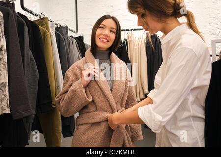 Jeune designer qui aide à essayer le manteau de la nouvelle collection à la jolie fille asiatique dans la boutique de vêtements Banque D'Images