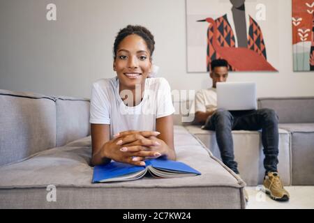 Jolie fille afro-américaine souriante, couchée sur un canapé avec un livre, qui regarde avec joie l'appareil photo tandis que l'ami travaille en arrière-plan sur un ordinateur portable à la maison Banque D'Images