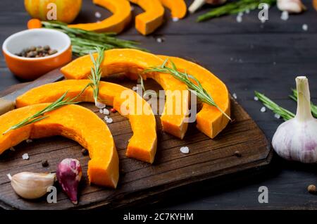morceaux de citrouille crus sur une planche à découper avec romarin, sel, épices sur une table sombre ancienne. Légumes préparés pour cuire au four. Automne cuisine maison Banque D'Images