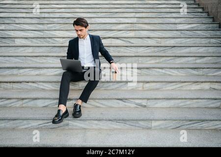 Jeune homme de brunes à barbe en chemise blanche et costume classique avec écouteurs sans fil assis dans les escaliers en extérieur et travaillant avec soin Banque D'Images