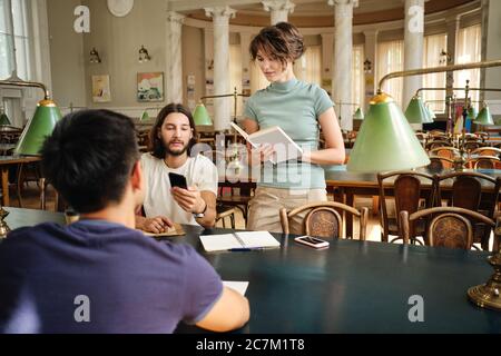 Groupe de jeunes étudiants occasionnels attrayants utilisant le téléphone portable tout en étudiant ensemble dans la bibliothèque de l'université Banque D'Images
