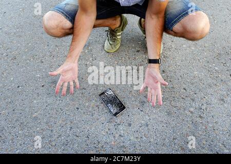 guy a laissé tomber son téléphone portable sur l'asphalte et l'a cassé. Un homme frustré avec un téléphone cellulaire cassé. L'homme est assis et tient un téléphone intelligent cassé Banque D'Images