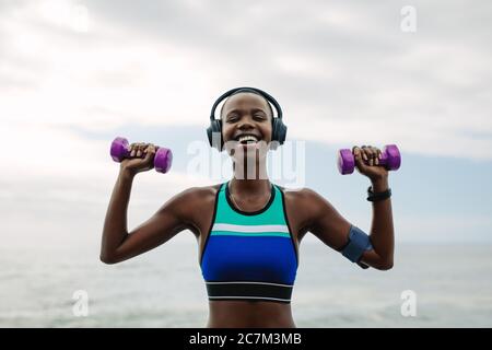 Femme heureuse portant des écouteurs s'exerçant avec des haltères à l'extérieur. femme de fitness portant des écouteurs faisant de l'entraînement près de la plage le matin. Banque D'Images
