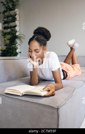 Jolie fille afro-américaine pensive couchée sur un canapé, lecture rêveur livre à la maison Banque D'Images