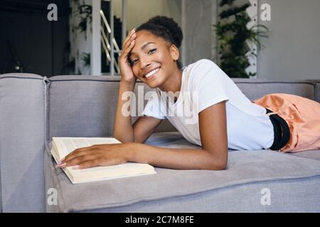 Jolie fille afro-américaine gaie allongée sur un canapé avec livre regardant avec plaisir dans l'appareil photo à la maison Banque D'Images
