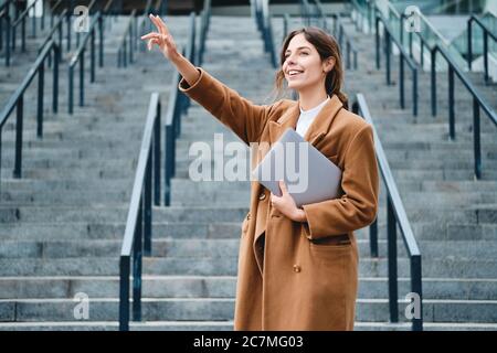 Jeune femme d'affaires souriante sous un manteau avec ordinateur portable agitant la main en plein air Banque D'Images