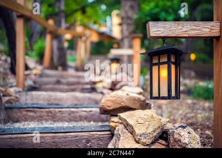 Lampe de lanterne suspendue illuminée en soirée sur poteau en bois dans le jardin japonais avec marches escaliers et feuillage vert de forêt par fond de balustrade Banque D'Images