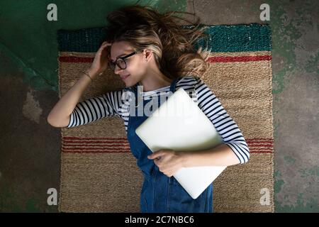 Vue du dessus de la femme mignonne en lunettes porter une combinaison bleue de jeans, couché sur un tapis de roseau sur le sol en béton, tenant l'ordinateur portable et se reposant après le travail. Banque D'Images