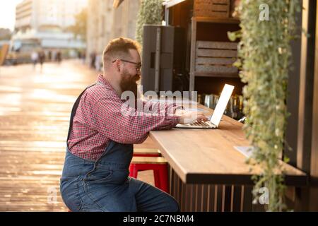 Portrait d'un boxeur hippster freelance à barbes, portant une combinaison bleue et une chemise à carreaux, travaillant sur un ordinateur portable assis dans un café/restaurant à l'extérieur. Côté Banque D'Images