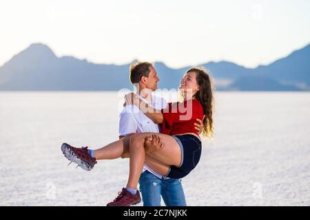 Jeune couple romantique heureux avec homme tenant une femme dans les bras sur blanc Bonneville Salt Flats près de Salt Lake City, Utah et vue sur les montagnes Banque D'Images