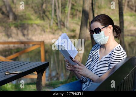 Femme assise sur un banc en bois dans un parc de lecture un livre tout en portant un masque chirurgical Banque D'Images