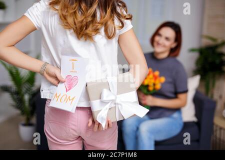 concept de fête des mères ou d'anniversaire - fille faisant la surprise pour la mère tenant une boîte cadeau et une carte de voeux faite à la main derrière le dos Banque D'Images