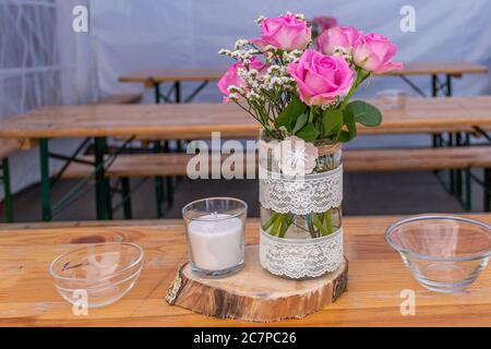 Bouquet de roses roses roses et petites fleurs blanches dans un vase en verre décoré de rubans blancs près d'une bougie dans un récipient en verre sur une base en bois Banque D'Images