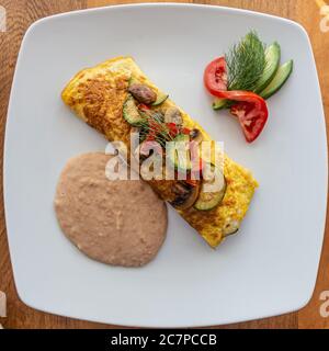 Omelette délicieuse au fromage, aux champignons et aux courgettes servie avec des haricots refrits et une petite salade d'avocat, de tomate et garnie d'un brin aromatique Banque D'Images