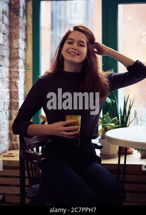 Belle sérieuse élégante et tendance, chic fille se trouve près de la fenêtre dans le café, sourires magnifiquement et boissons saines smoothie végétalien ou latte. Charmant Banque D'Images