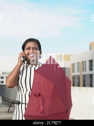 Femme d'affaires indienne parlant au téléphone et tenant un parapluie rouge. Extérieur. Banque D'Images