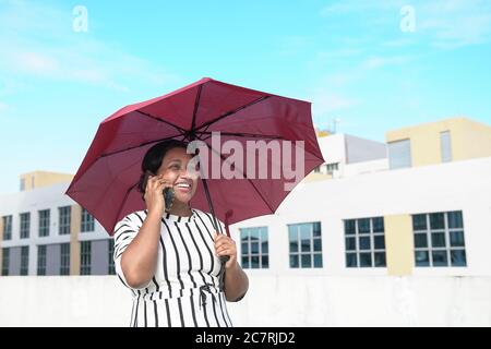 Femme d'affaires indienne souriante parlant au téléphone et tenant un parapluie rouge. Fond urbain. Banque D'Images