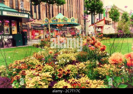 Vue à travers les fleurs d'été colorées : Corneliusplatz dans le centre-ville de Düsseldorf avec un kiosque à journaux et un carrousel nostalgique. Filtre rétro. Banque D'Images