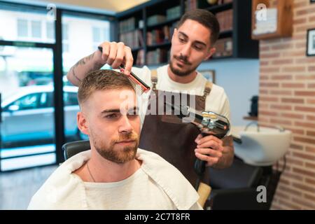 Coiffeur jeune peignant et sécher les cheveux du client mâle dans un barbershop Banque D'Images