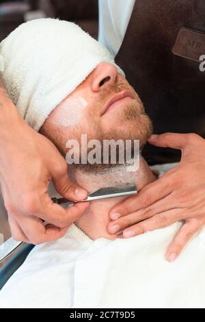 Photo verticale d'un coiffeur se raser la barbe d'un jeune client dans une serviette avec une lame Banque D'Images